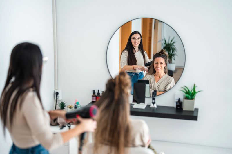 Young adult Caucasian hairdresser in head spa salon skillfully blow drying client's hair with mirror reflection showing both smiling. Modern salon setup with beauty tools on counter.