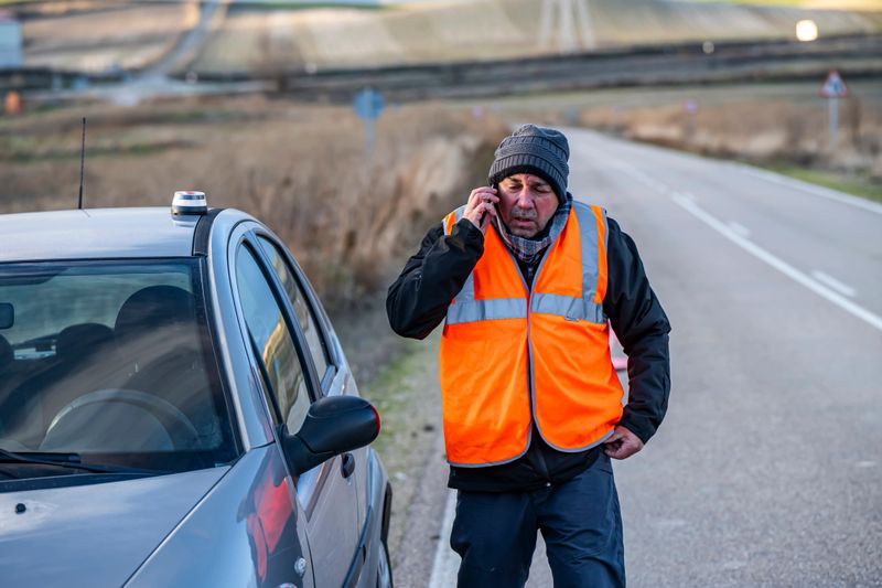 Man outside his vehicle contacting emergency services and roadside assistance with a tow truck. His vehicle has broken down and he is using the mandatory V16 warning light on the vehicle's dashboard, ready to be placed on the roof.