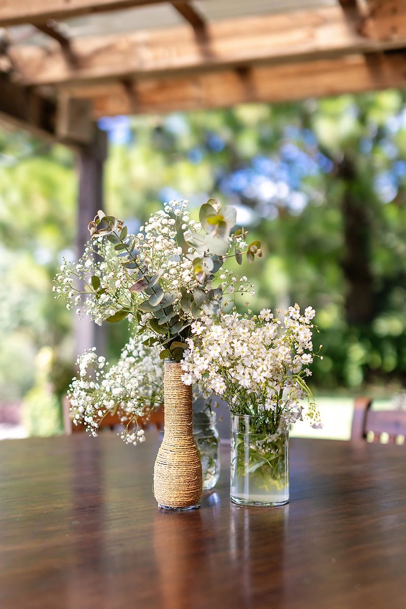 Elegant floral arrangement with white gypsophila and eucalyptus in glass vases on a wooden table. Natural wedding decor with soft light and blurred garden background.