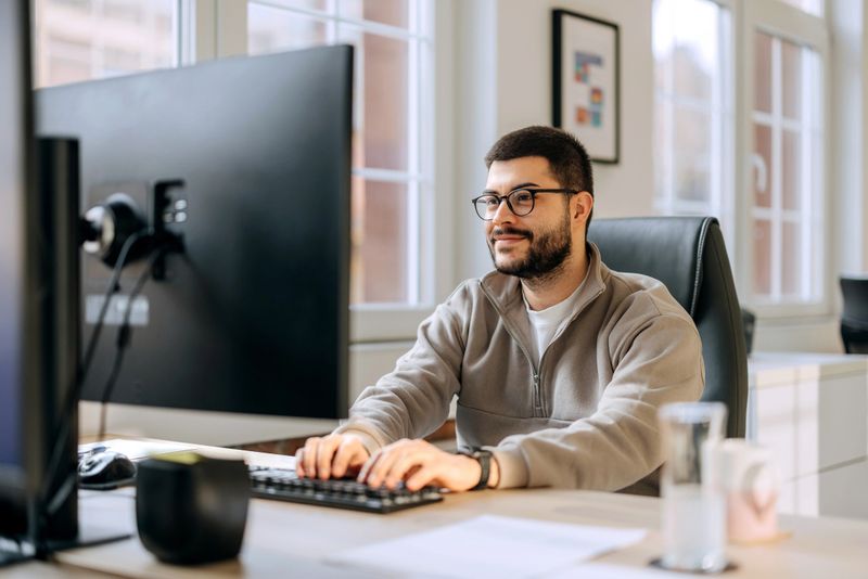 A casually clothed professional man focused on his work while using a desktop PC in a modern office environment. The scene represents everyday office life, productivity, and a relaxed business atmosphere.