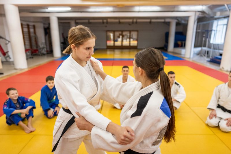 Two female judo students demonstrate a grip during a training session on a bright yellow and red tatami, while peers in blue and white judogi observe in a spacious dojo.