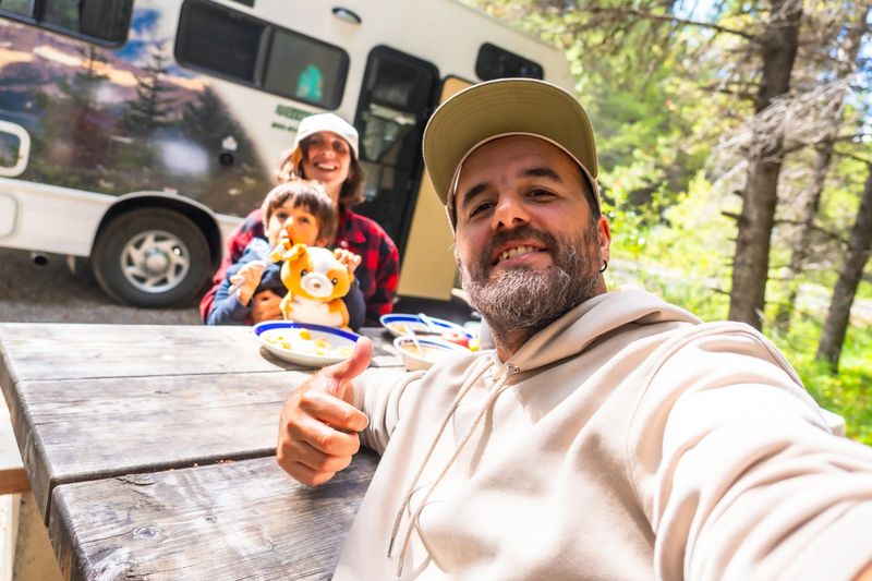 Happy family taking a selfie while enjoying a meal at a picnic table outside their campervan in the beautiful banff national park, canadian rockies