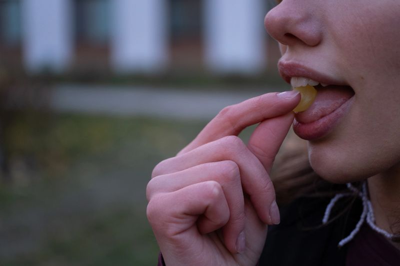 Detailed close-up of female lips and fingers as she places a yellow throat lozenge onto her tongue. The image represents cold and flu care and daily personal health routine in an urban environment. Suitable for pharmacy promotion, medical articles, healthcare education, wellbeing content, and seasonal illness campaigns.