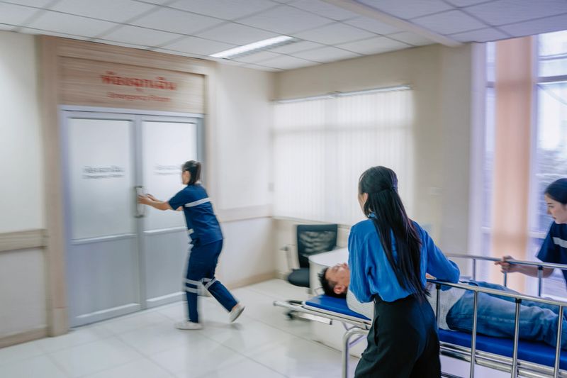 Asian male and female healthcare workers urgently push a male patient on a stretcher through hospital doors and into an emergency treatment area. Filmed in a realistic handheld style, the scene highlights teamwork, rapid response, and the decisive moments of hospital emergency care. Bangkok