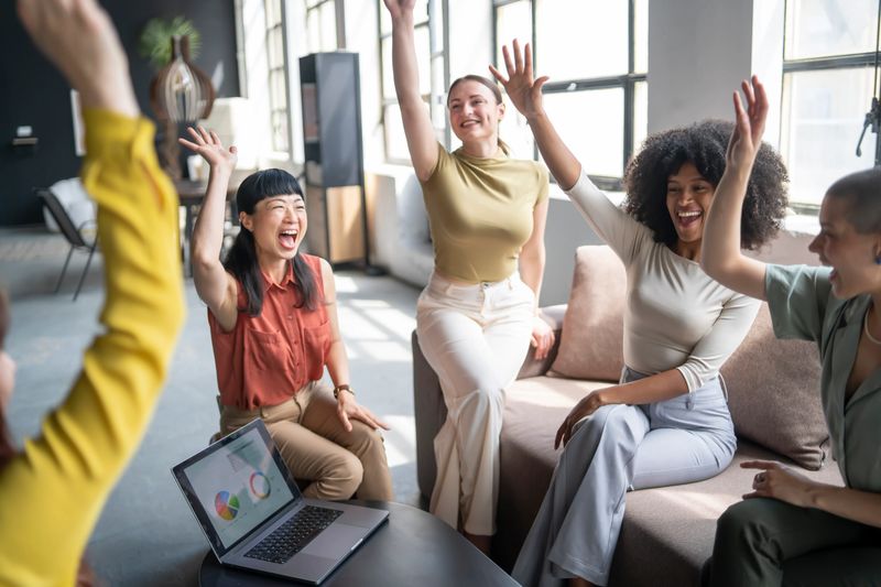 Group of diverse businesswomen raising hands in celebration, happy about positive results shown on laptop, enjoying teamwork and success in modern office
