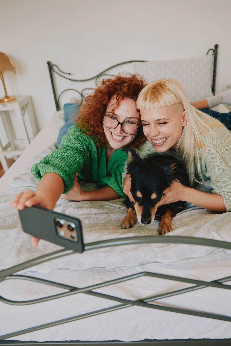 Two adult women share a playful moment on a bed with their dog, posing for a selfie in a bright, cozy apartment. A warm, friendly scene perfect for lifestyle and LGBTQ themes.