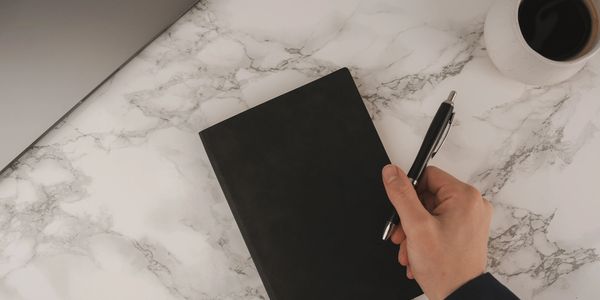 Hand holding pen over black notebook on a marble table with laptop and coffee.