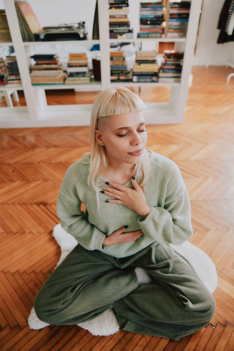 Young hipster woman sits in yoga pose while meditating in bright living room of apartment with bookshelf and plate of fruit.
