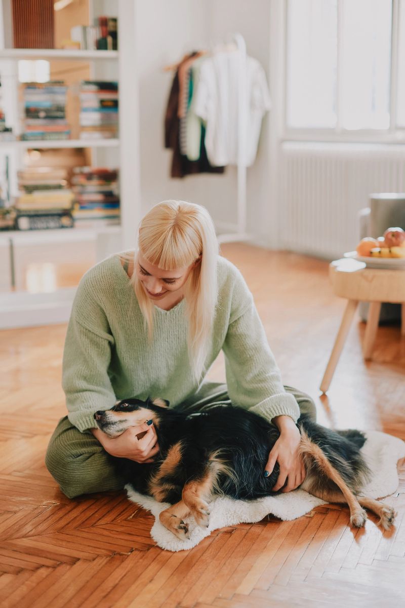 A warm indoor scene shows an adult woman relaxing with her dog on a polished wooden floor in a bright living room. Soft light and cozy furniture create a homely, comforting vibe.