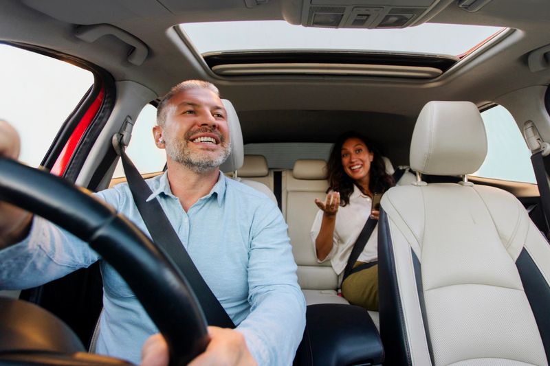 Nice ride. Happy male driver riding car, looking at lady sitting in auto on back passenger seat, woman talking with man, windshield view