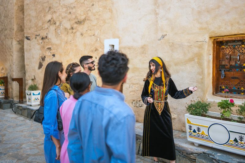 A young Middle Eastern female guide in traditional Saudi dress is leading a group of diverse tourists at Abu Sarrah Palace. She is gesturing and explaining, while the tourists are attentively listening and observing the historic architecture.