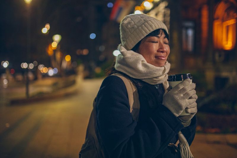 A smiling woman walks through the city at night holding a takeaway coffee, enjoying a calm urban evening.