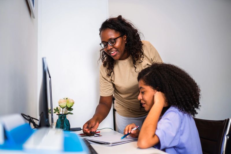 A Black woman wearing glasses and a tan t-shirt stands next to a young girl with curly hair, helping her with her studies. They are at a white desk with a computer monitor and a notebook. The woman is pointing at the notebook while the girl holds a pen, both focused on the task. The scene depicts a supportive educational environment at home.