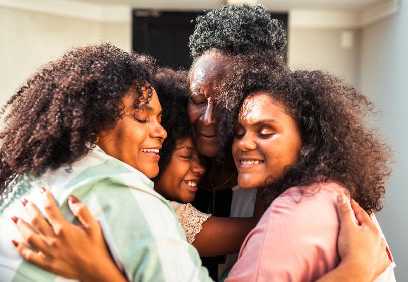 A heartwarming close-up shot of four multi-generational Black women, including a grandmother, mother, and two younger women, embracing each other tightly. Their eyes are closed in contentment, and wide smiles light up their faces, conveying deep love, happiness, and strong family connection. The image captures a moment of genuine affection and joy among family members.