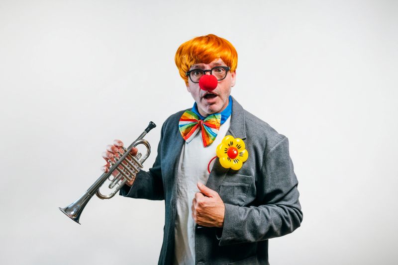 Clown performer with red nose, orange wig, and colorful bow tie holding a trumpet