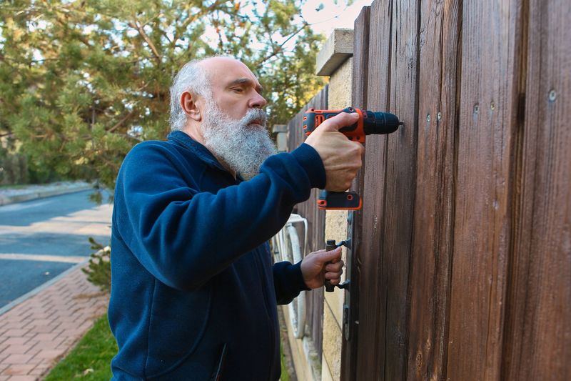 Senior man drilling wooden fence with determined focus during home repair. Concept of DIY maintenance, outdoor renovation tasks, household improvement, and handyman visuals.