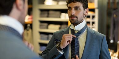 Man adjusting his tie in front of a mirror wearing a blue suit.