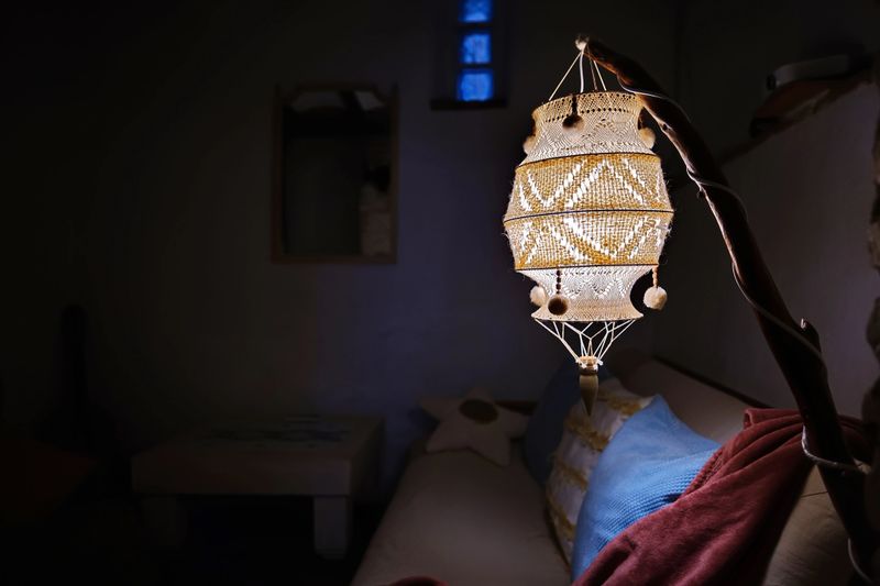 A decorative hanging lantern glows softly in a dim living space, featuring intricate beadwork and woven patterns that create a warm, inviting atmosphere among cushions and textiles.