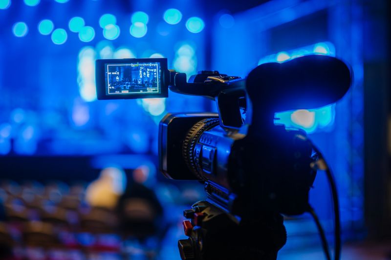 Video camera on tripod is focused on live performance stage illuminated by blue lights, emphasizing the dynamic atmosphere of event recording