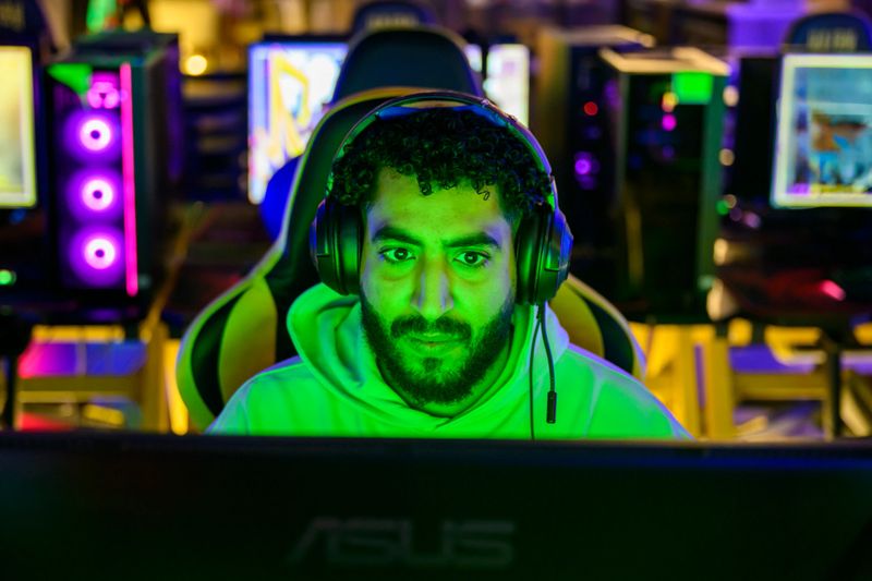 Focused young Saudi adult man wearing gaming headphones and a green hoodie, concentrating on a computer screen in a dimly lit room with vibrant RGB lighting, immersed in an online gaming session