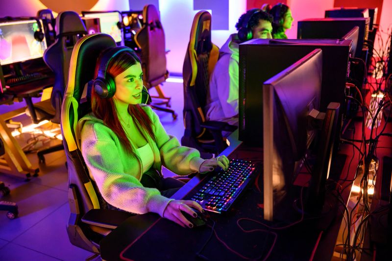 Young woman in headphones intently gaming on a PC with glowing keyboard and mouse, focused on the screen in a dim, neon-lit esports arena during online competition