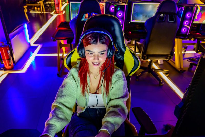 Young adult woman with headset and microphone, sitting in a gaming chair, smiling and focused while streaming or competing in a neon-lit dark room with monitors glowing