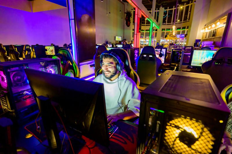 Smiling young adult gamer wearing a headset and focused at a brightly lit RGB desktop workstation in a modern gaming cafe, playing and streaming a competitive online video game with joy