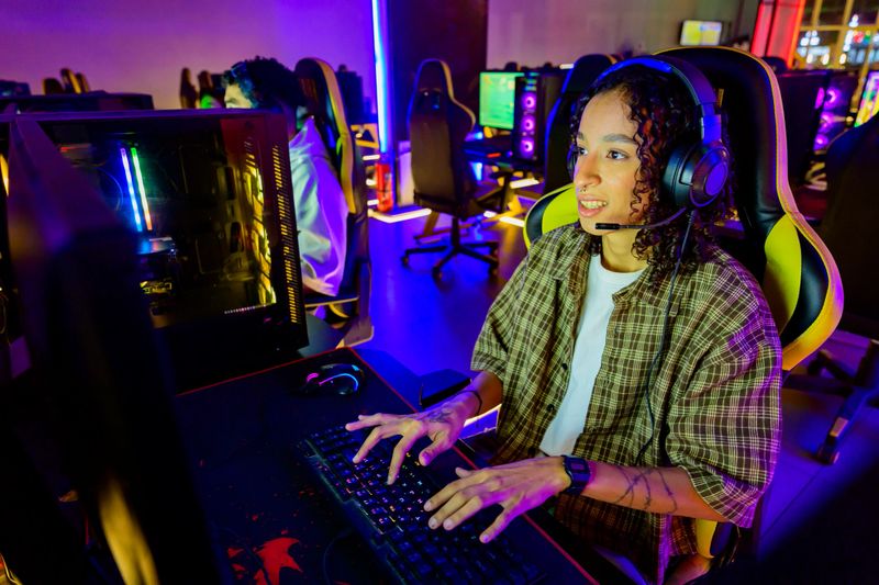 Young adult woman gaming online in an internet cafe, wearing a headset with a microphone, focusing on the screen and typing on the keyboard during an esports competition