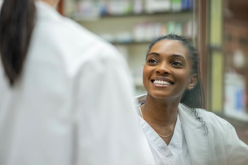 A friendly Black woman in a white lab coat smiles while speaking with a colleague in a pharmacy or clinical setting. The scene conveys teamwork, professionalism, and positive healthcare collaboration.