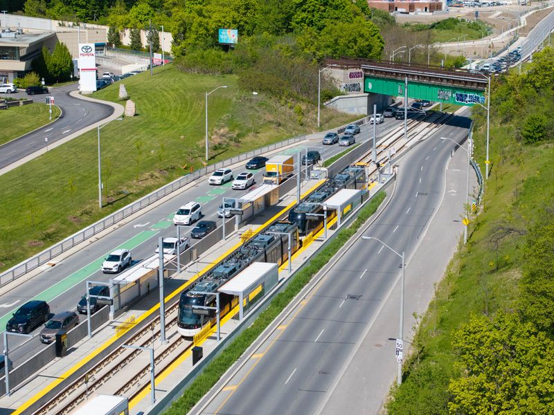 A high-angle drone shot capturing the new Eglinton Crosstown Light Rail Transit (LRT) vehicles traveling through the scenic intersection of Eglinton Avenue East and Leslie Street in Toronto. The image showcases the sleek, modern architecture of residential towers (Auberge on the Park) juxtaposed against the lush greenery of Sunnybrook Park and Wilket Creek. This photo represents urban development, modern public transportation, and sustainable city living in Canada's largest metropolis.