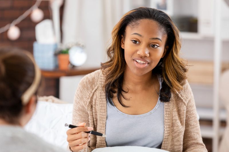 Two women in a counseling session, one speaking while holding a pen, indoors.