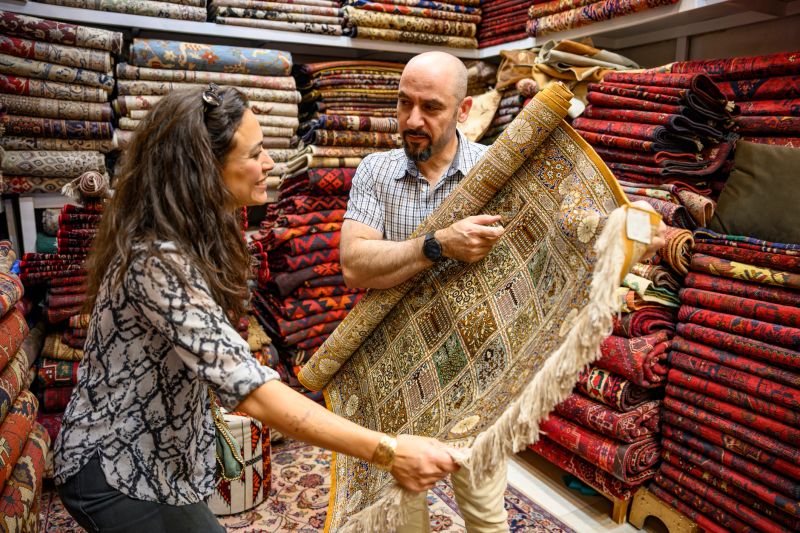 Customer and merchant inspecting a traditional handmade carpet, surrounded by stacks of colorful textiles and authentic rugs, highlighting craftsmanship and local shopping experience