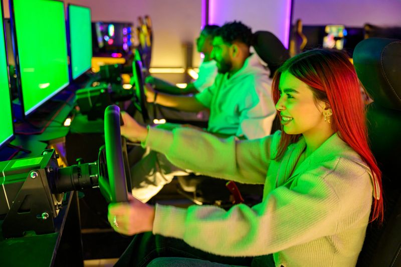 Young woman smiling and focused while racing on a steering wheel and pedal simulator, competing with friends in a vibrant neon-lit esports arena full of energy and excitement