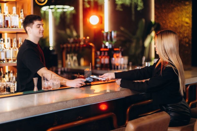 A young woman making a cashless payment to a bartender at a stylish, well-lit bar. The lively atmosphere of the bar emphasizes hospitality, interaction, and a modern lifestyle.