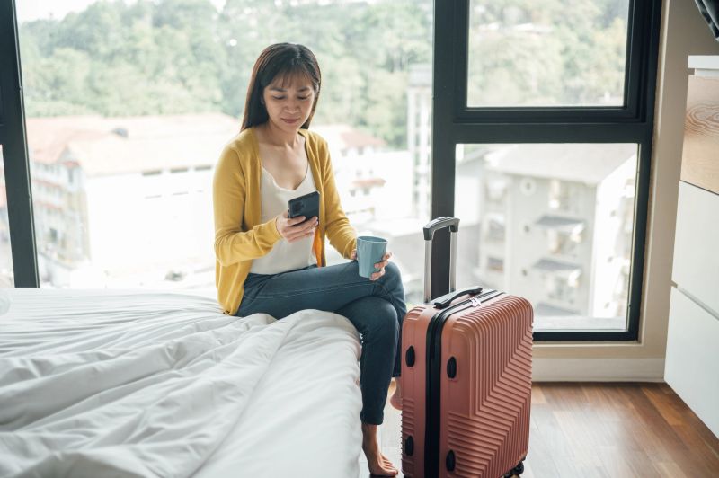 A female tourist sits on a hotel bed with her luggage, using a mobile phone to check local maps or travel bookings