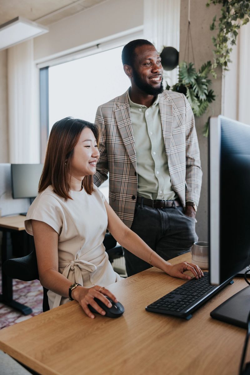 Two professionals collaborate in a modern office, one seated at a wooden desk using a mouse and keyboard while the other stands beside, smiling as they review data on a large computer monitor.