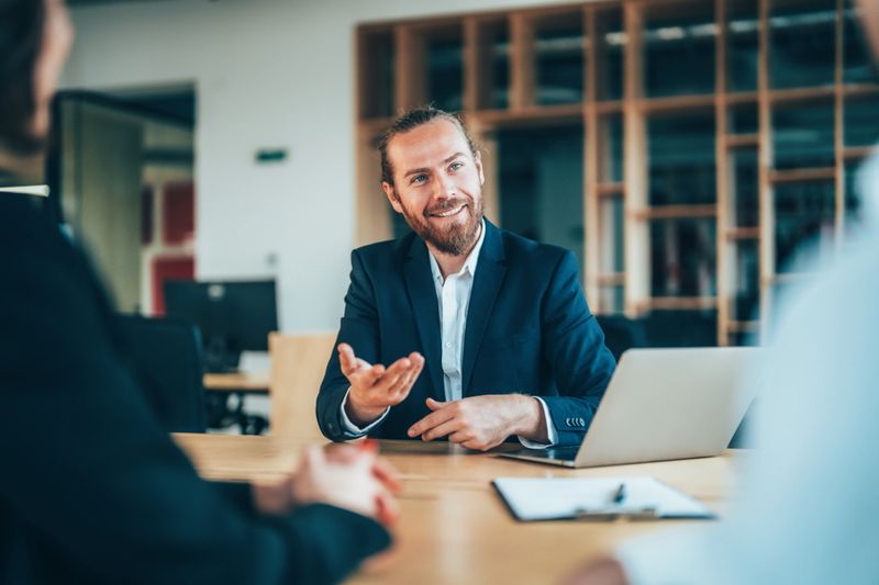 Shot of confident business persons sitting on a desk in the office and sharing ideas. Businessman and businesswoman in meeting using laptop and discussing business strategy and supply chain.