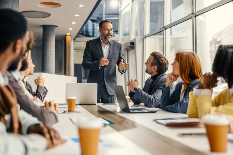 Mature male professional in a suit leads a diverse team around a modern conference table, presenting and facilitating strategic discussion, collaboration and attentive teamwork