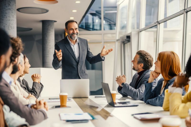 Confident business leader standing at a table, explaining a strategy to diverse colleagues listening attentively during a corporate meeting in a modern office