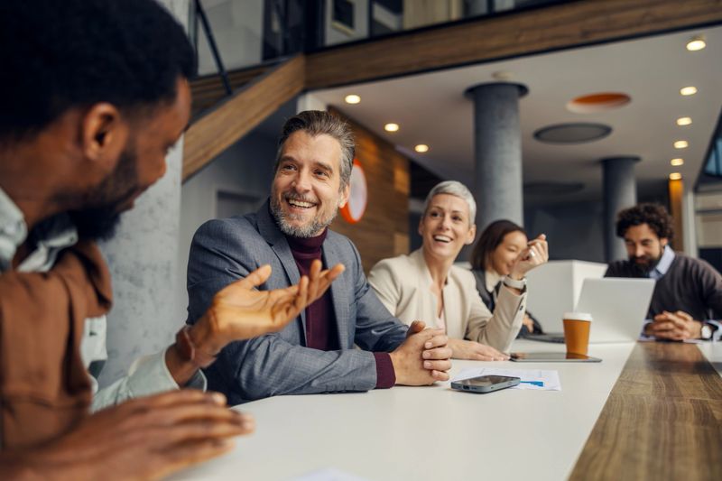 Diverse business professionals smiling and communicating during a cheerful meeting in a modern office, fostering teamwork, collaboration, and productive conversation