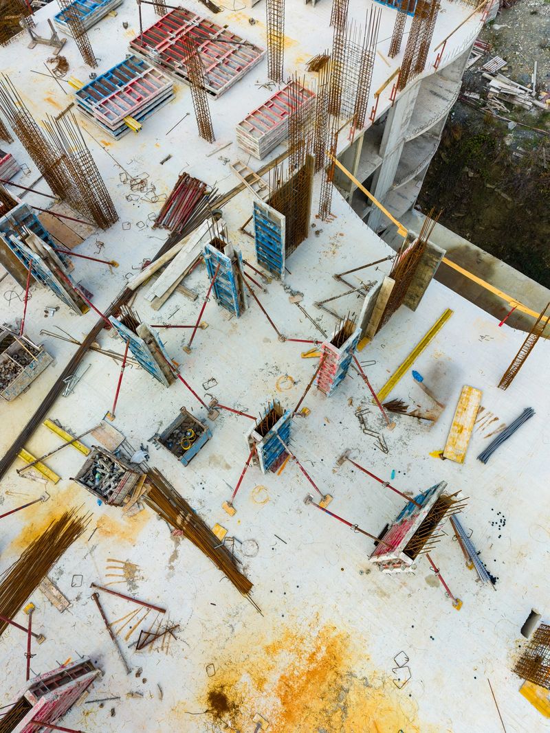 Workers are busy at a construction site. Steel beams are arranged around the area. Equipment and tools are visible. The sun is shining brightly over the scene.