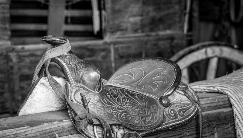 Black and white image of a western saddle resting in an old barn, with the saddle horn prominently visible. The photograph emphasizes rustic textures, traditional craftsmanship, and a timeless rural atmosphere, evoking heritage, nostalgia, and classic western culture.