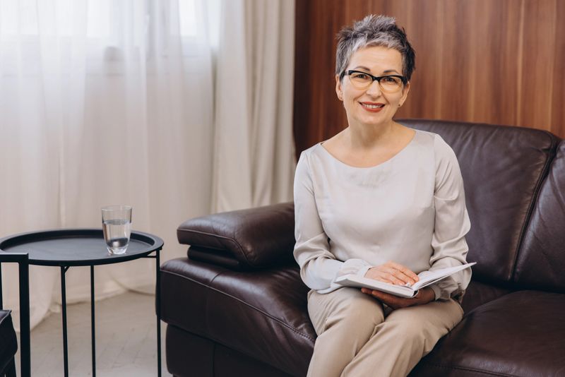 Professional woman psychologist or therapist smiling, sitting on a leather sofa in a comfortable office setting, holding a book