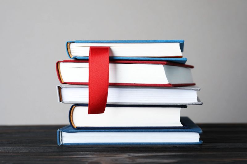 Book with red ribbon bookmark and colorful ones on wooden table against grey background, closeup