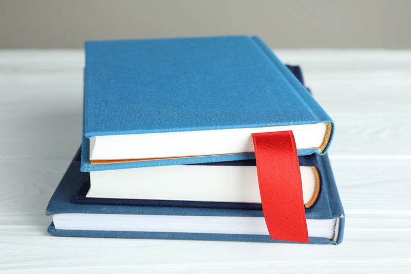 Book with red ribbon bookmark and colorful ones on white wooden table against grey background, closeup