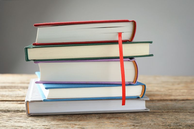 Book with red ribbon bookmark and colorful ones on wooden table against grey background, closeup
