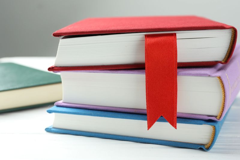 Book with red ribbon bookmark and colorful ones on white wooden table against grey background, closeup