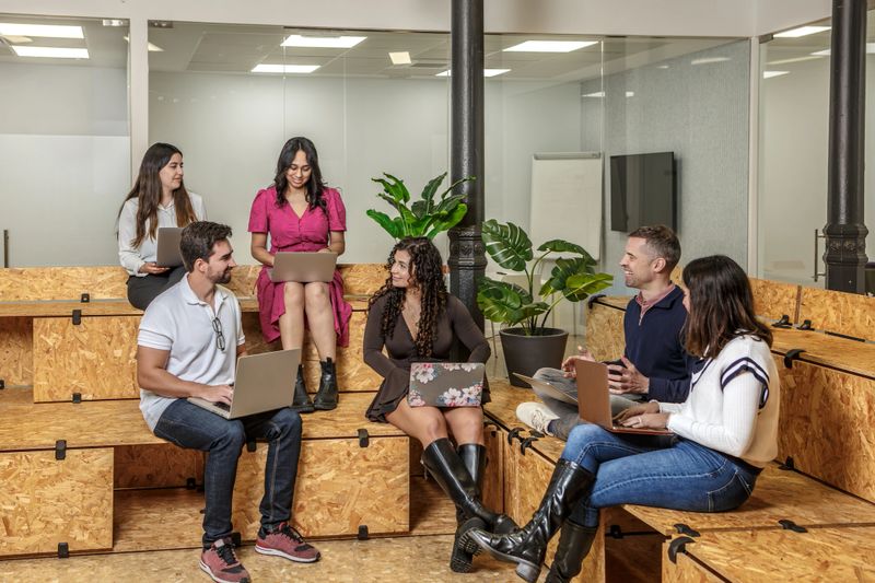 In a coworking space, young professionals collaborate on projects while sitting on wooden steps. The setting features modern decor and plants.