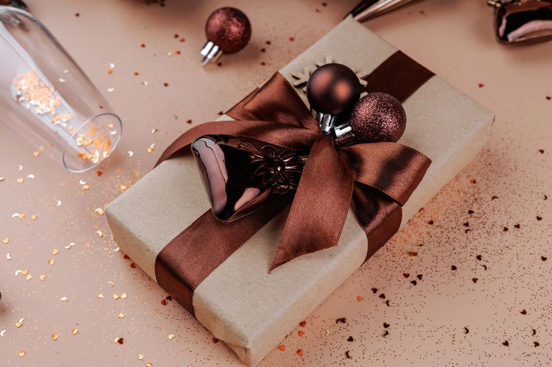 Close-up of gift box with bronze ribbon, holiday ornaments and confetti on beige background