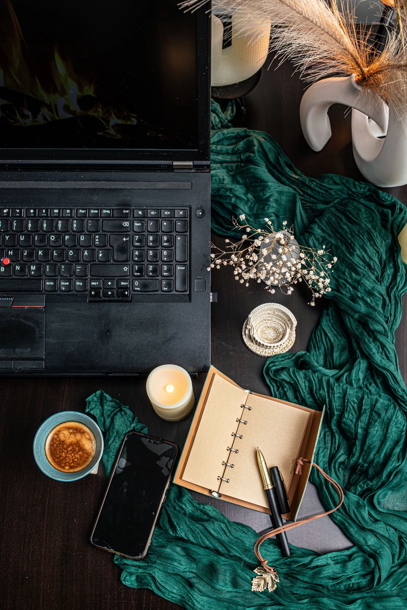 Cozy and atmospheric flat lay workspace with notebook, coffee cup, candles, smartphone, and warm ambient light. A minimalist to-do list for 2026 lies open on a dark desk, surrounded by soft green textile, dried flowers, and decorative elements. Concept of planning, productivity, slow living, mindfulness, self-care, home office, and New Year goals. Warm tones create a calm and inspirational mood.